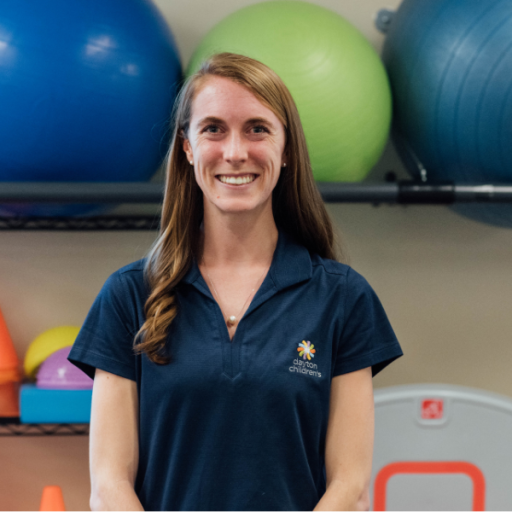 A smiling woman with long brown hair, wearing a dark blue polo shirt and a delicate necklace, stands in a brightly lit gym with exercise balls behind her.