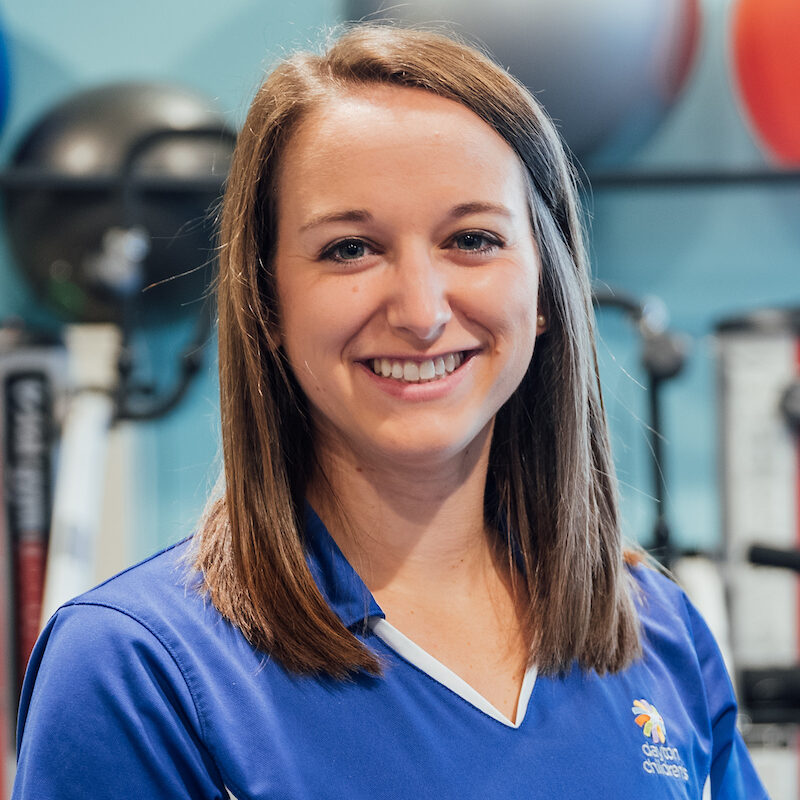 A professional headshot of a smiling woman with straight shoulder-length brown hair, wearing a blue and white athletic polo shirt in a gym setting.