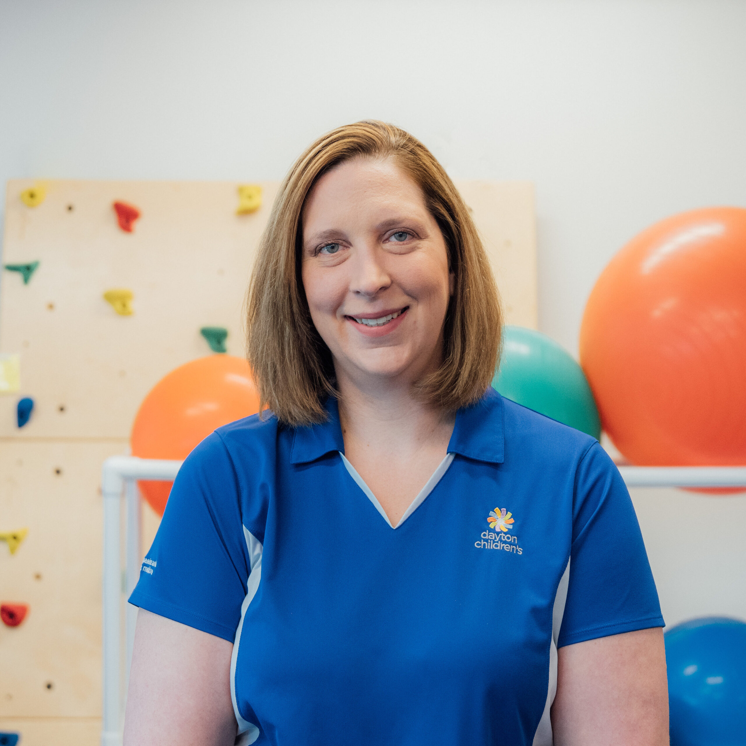 A friendly female healthcare professional with medium-length hair smiles in a blue polo shirt in a therapy gym, with exercise equipment behind her.